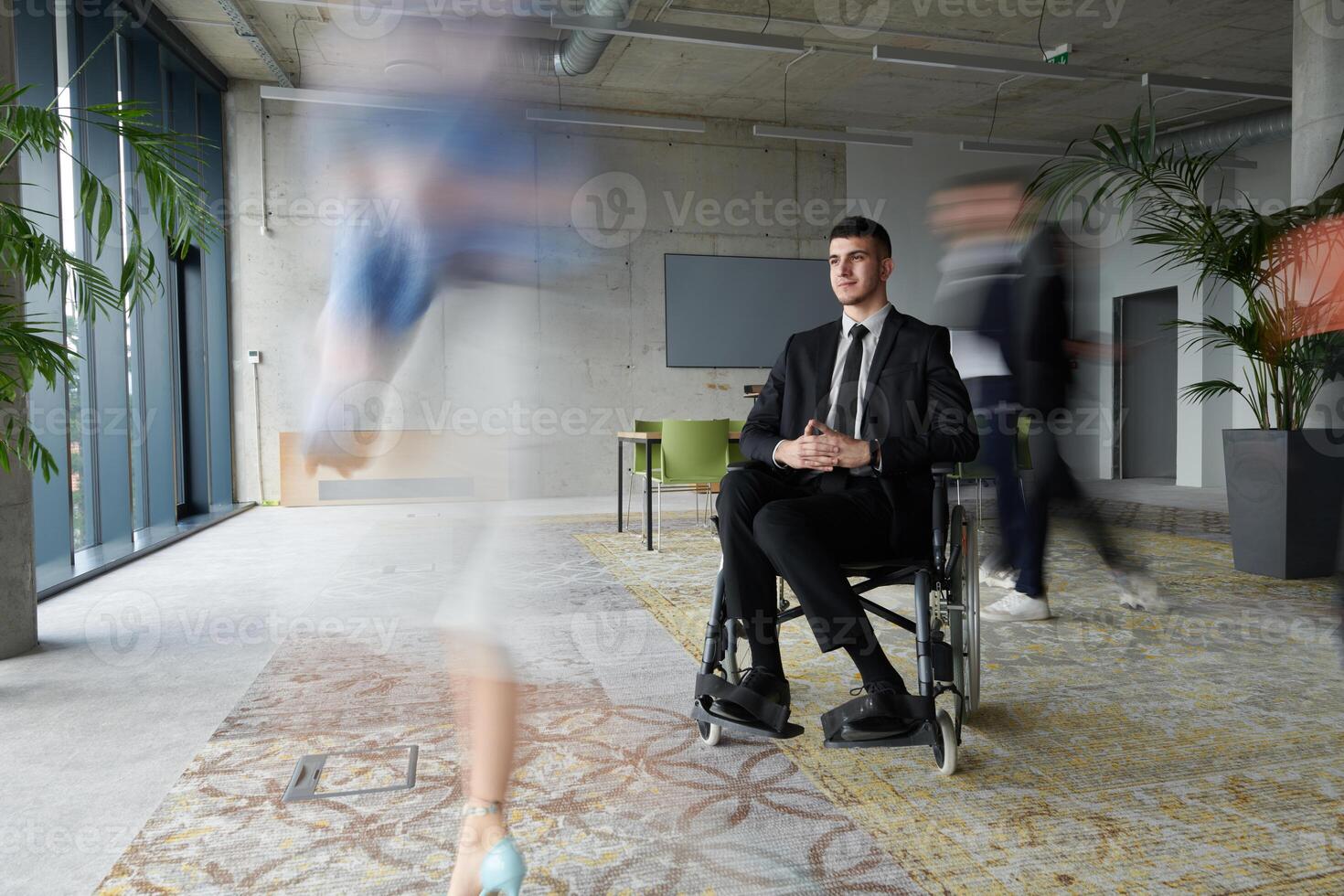 A businessman in a wheelchair navigating through a busy office, surrounded by his colleagues who are actively engaged in their work and collaboration photo