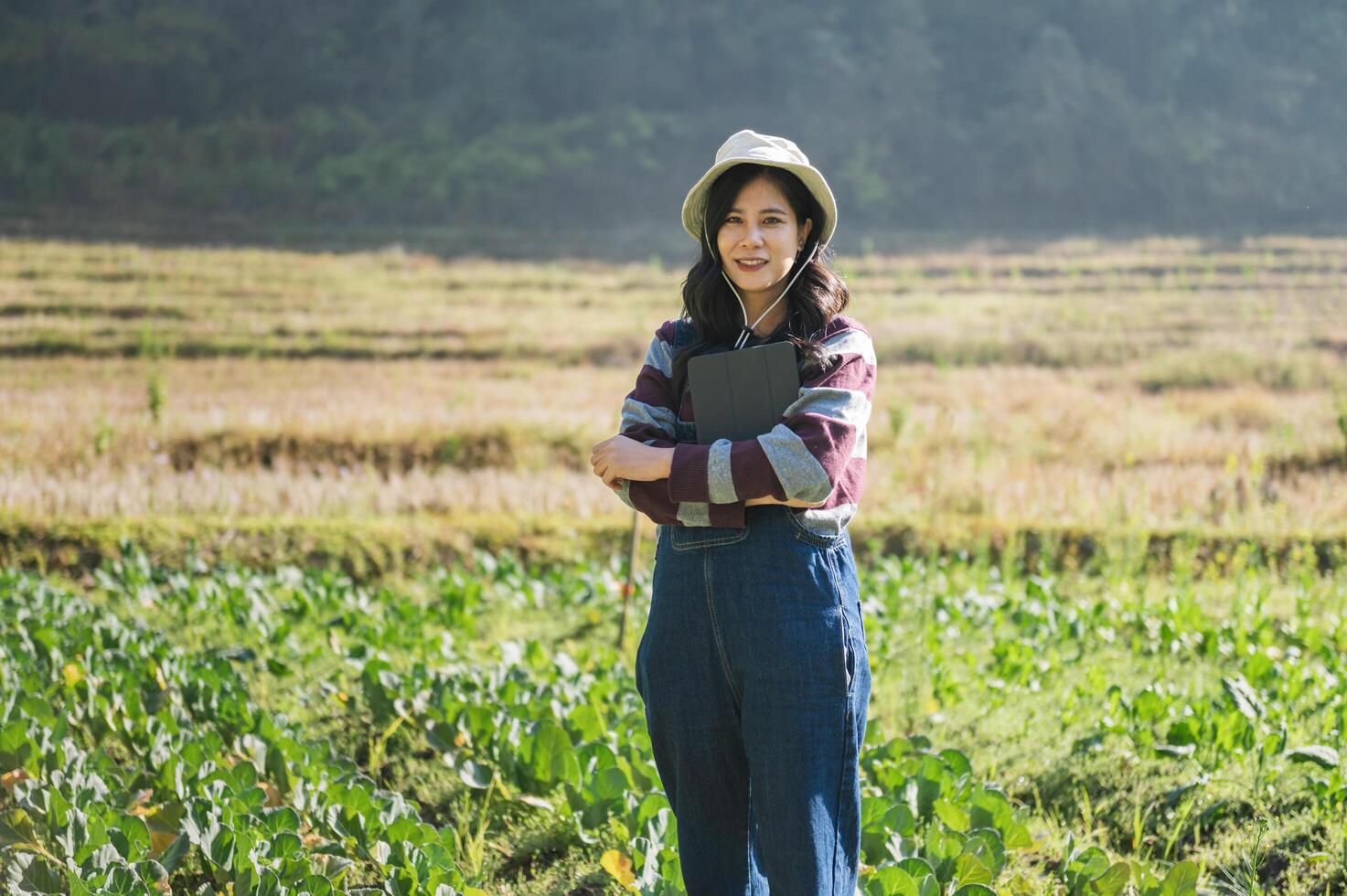 woman wearing a hat and blue overalls stands in a field of green plants photo