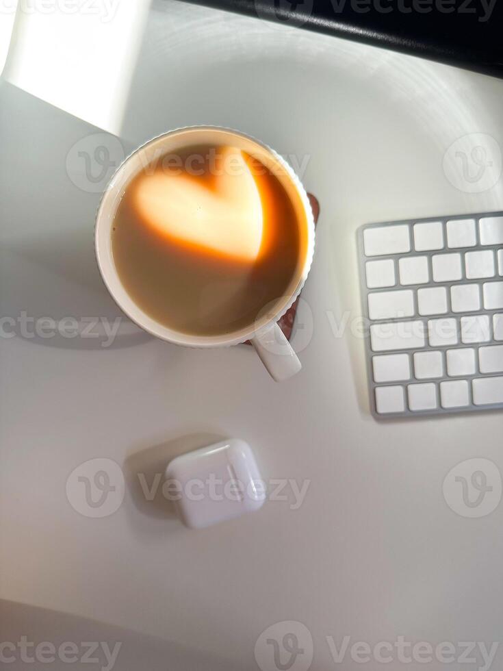 Cup of coffee with a heart-shaped reflection for Valentines day, placed on a clean white desk. Soft lighting and minimalistic workspace. Love and technologies concept. photo