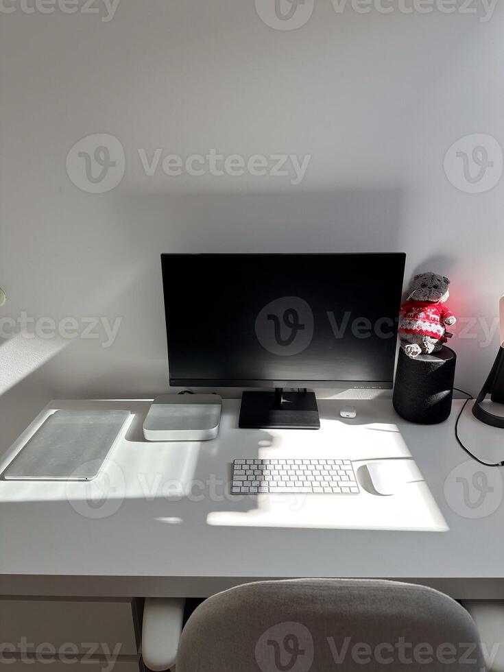 Minimalist white workspace with a computer monitor, keyboard, and other tech accessories on a clean desk. Bright sunlight streaming through the window highlights the neat and organized setting photo
