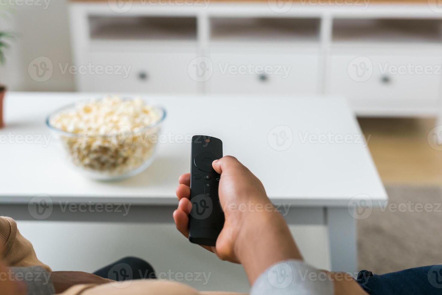 Hand holds a remote control while a bowl of popcorn sits on a white table in a modern living room. Relaxing and cozy atmosphere, perfect for watching movies or streaming content at home photo