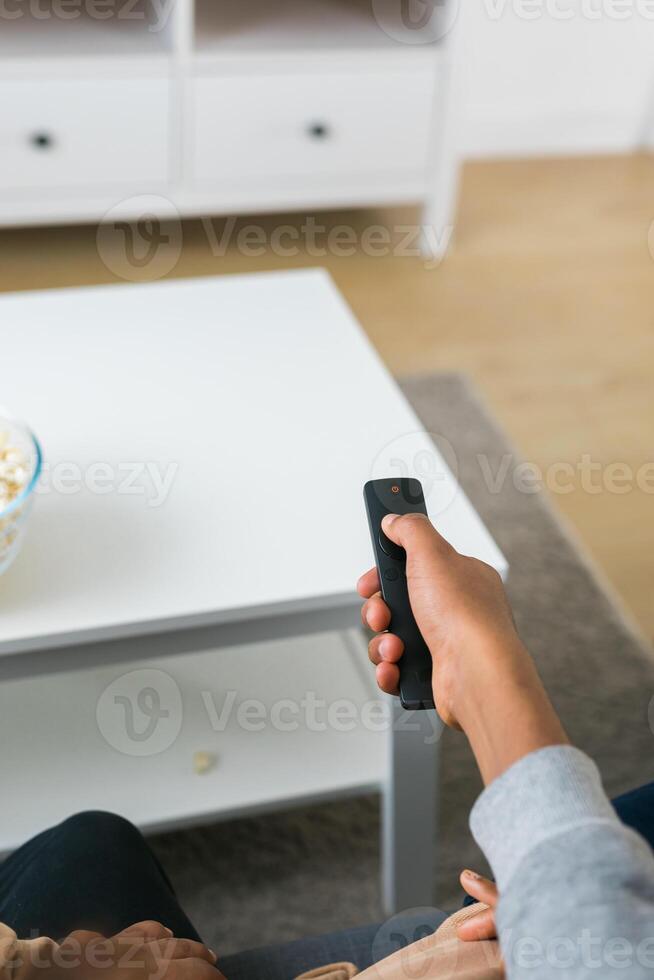 Hand holds a remote control while a bowl of popcorn sits on a white table in a modern living room. Relaxing and cozy atmosphere, perfect for watching movies or streaming content at home photo
