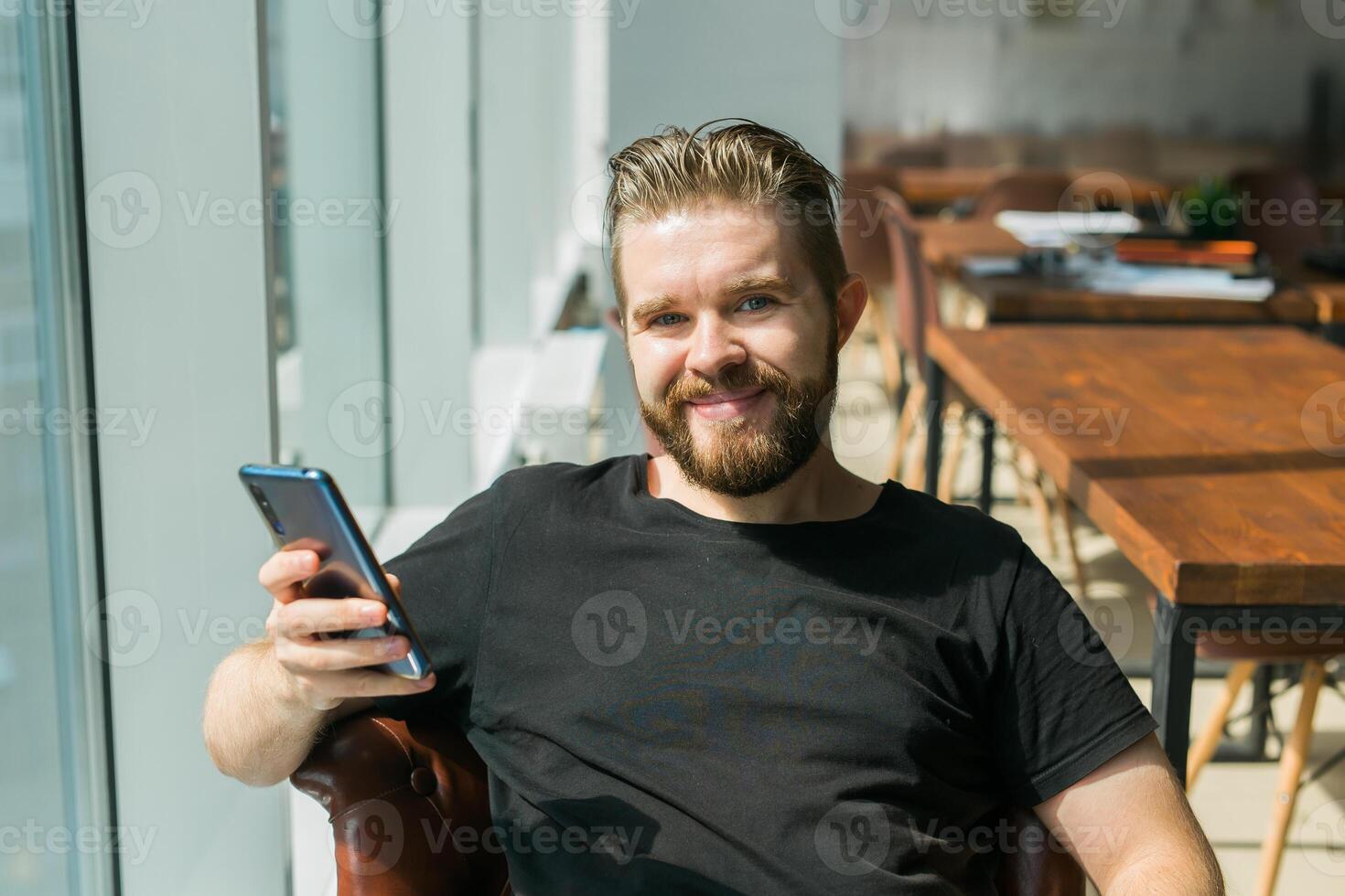 Smiling young adult freelance business man using mobile phone checking social media network feed or message chat sitting on chair. Online digital communication and gadget addiction concept photo