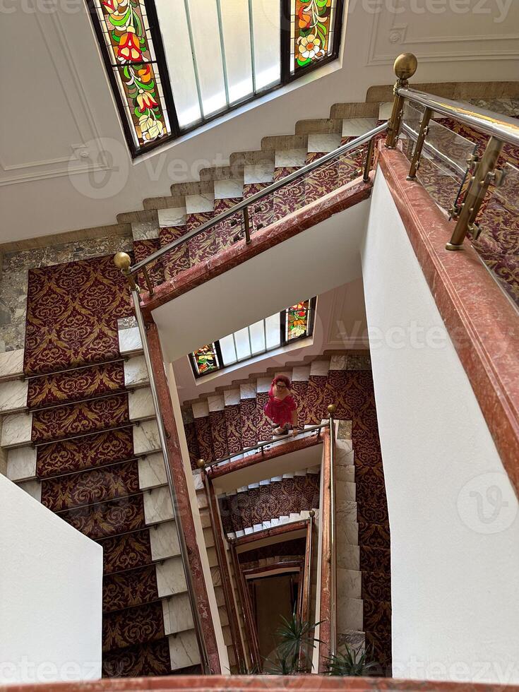Woman in a pink floral dress ascending a grand staircase adorned with intricate stained glass and ornate carpeting. Blend of timeless elegance and vibrant charm in a classic architectural setting photo