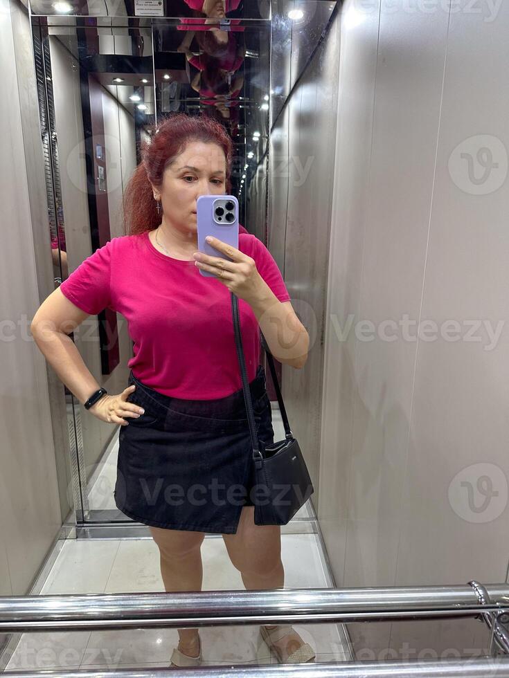 Millennial woman in a pink shirt taking a selfie in a mirrored elevator. Technologies and photo for social networks