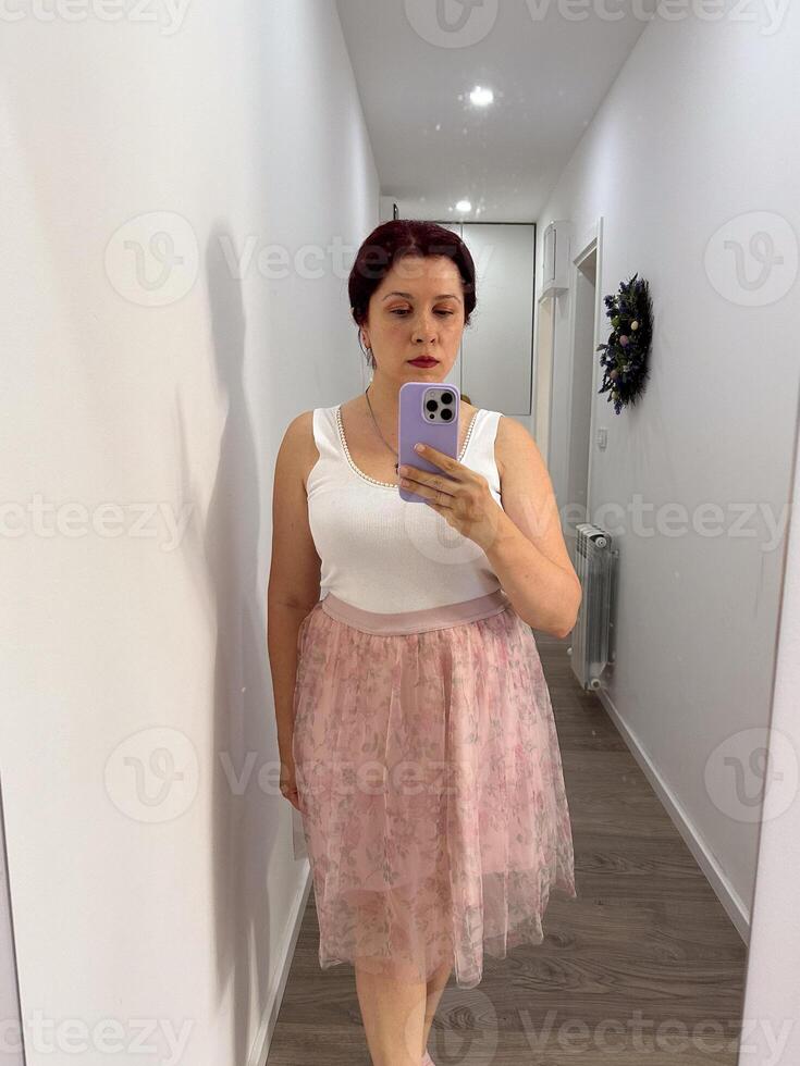 Woman takes a mirror selfie in a minimalist home hallway, wearing a white tank top and a pastel pink floral skirt. Soft, feminine and casual vibe ideal for a warm day or relaxed outing. photo