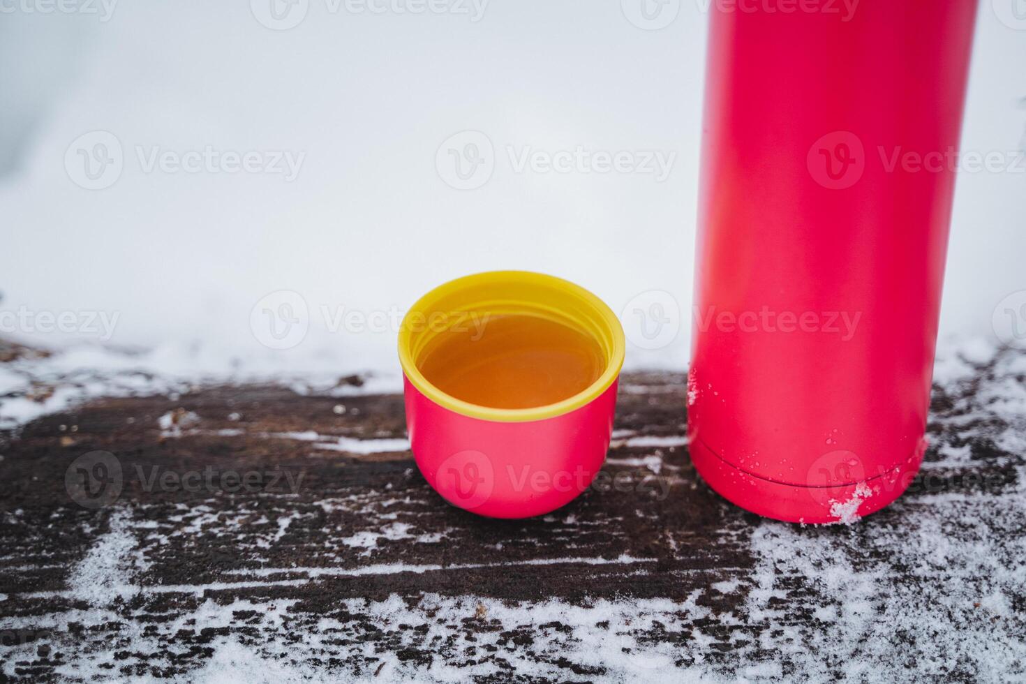 Magenta plastic cup with Dianhong tea and red thermos on snowy wooden table photo