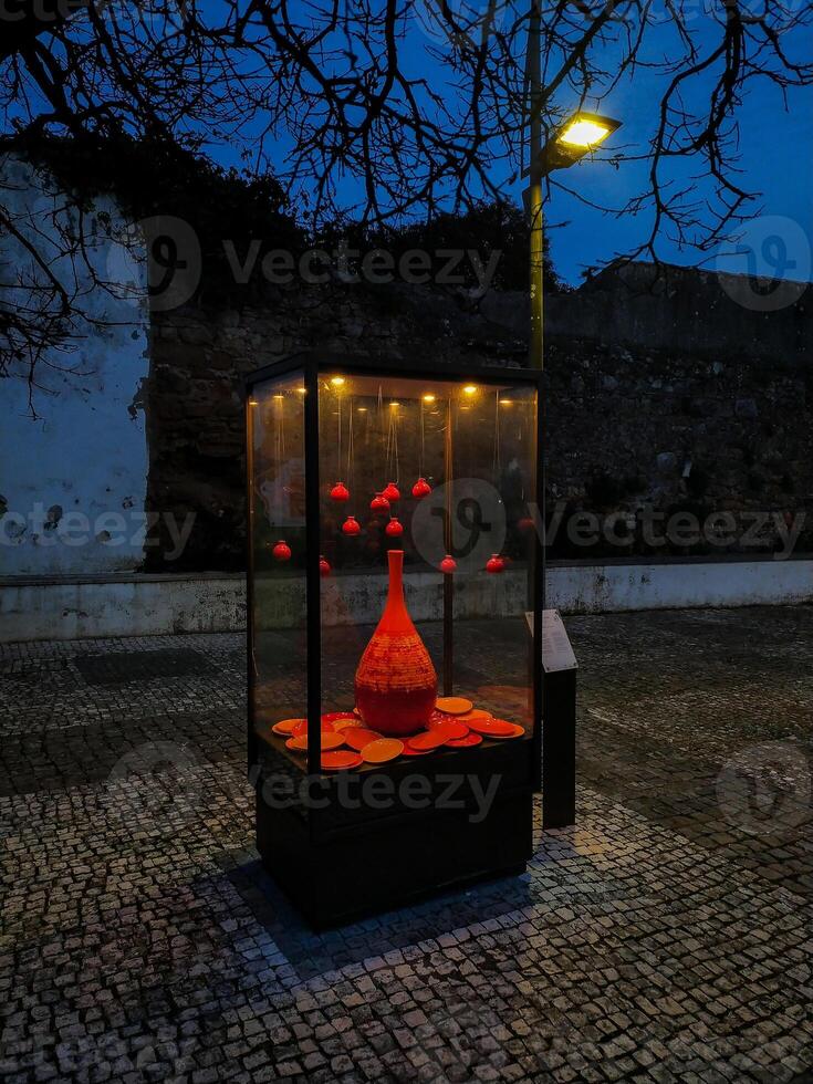 Dramatic nighttime shot of a glass display case showcasing a vibrant red ceramic vase and smaller hanging ornaments. The cobblestone setting and ambient lighting create a captivating, artistic mood. photo