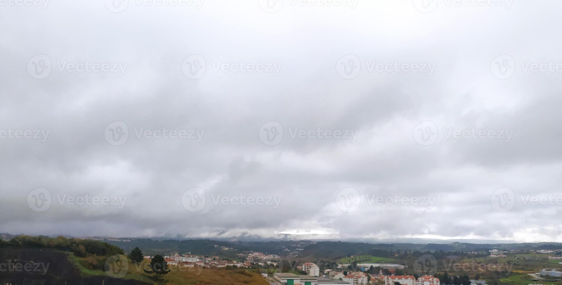 Serene landscape photo showcasing a sprawling town nestled amongst rolling hills under a dramatic, overcast sky. Ideal for travel, nature, or real estate projects needing a moody atmosphere.