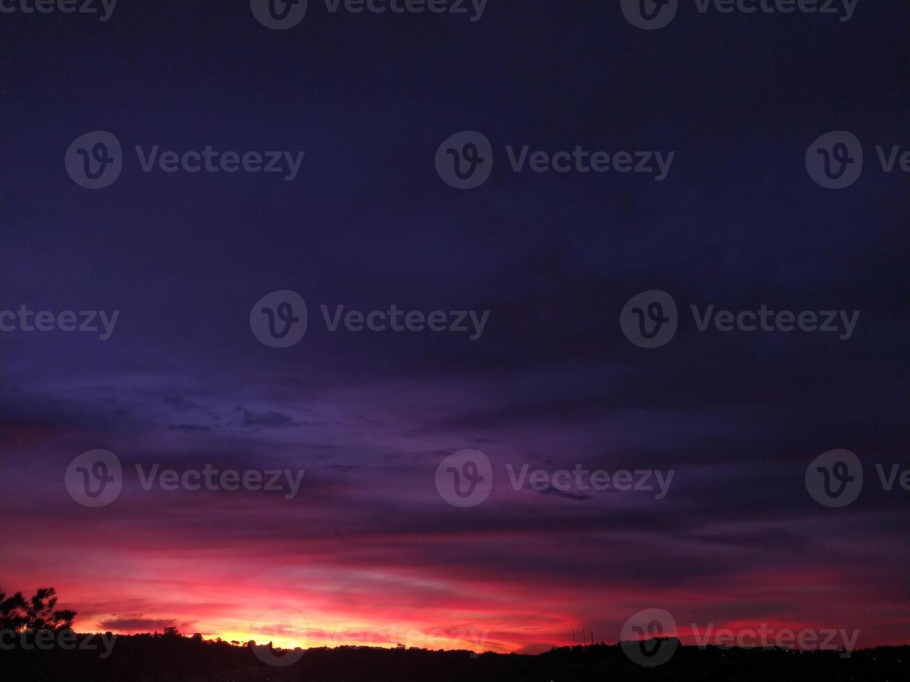 Dramatic sunset landscape showcasing vibrant crimson and deep purple hues across the sky. Silhouetted trees and distant cityscape add depth. Ideal for travel, nature, and atmospheric backgrounds. photo
