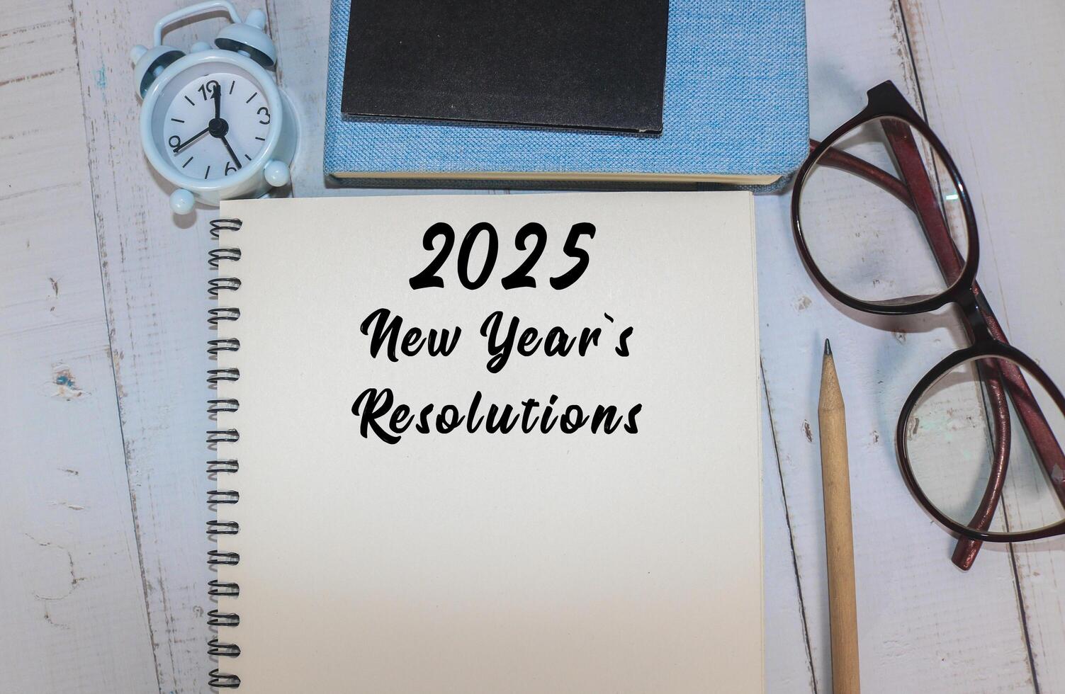 A workspace featuring a notepad with 2025 New Year's resolutions, glasses, a pen, books, and clock photo
