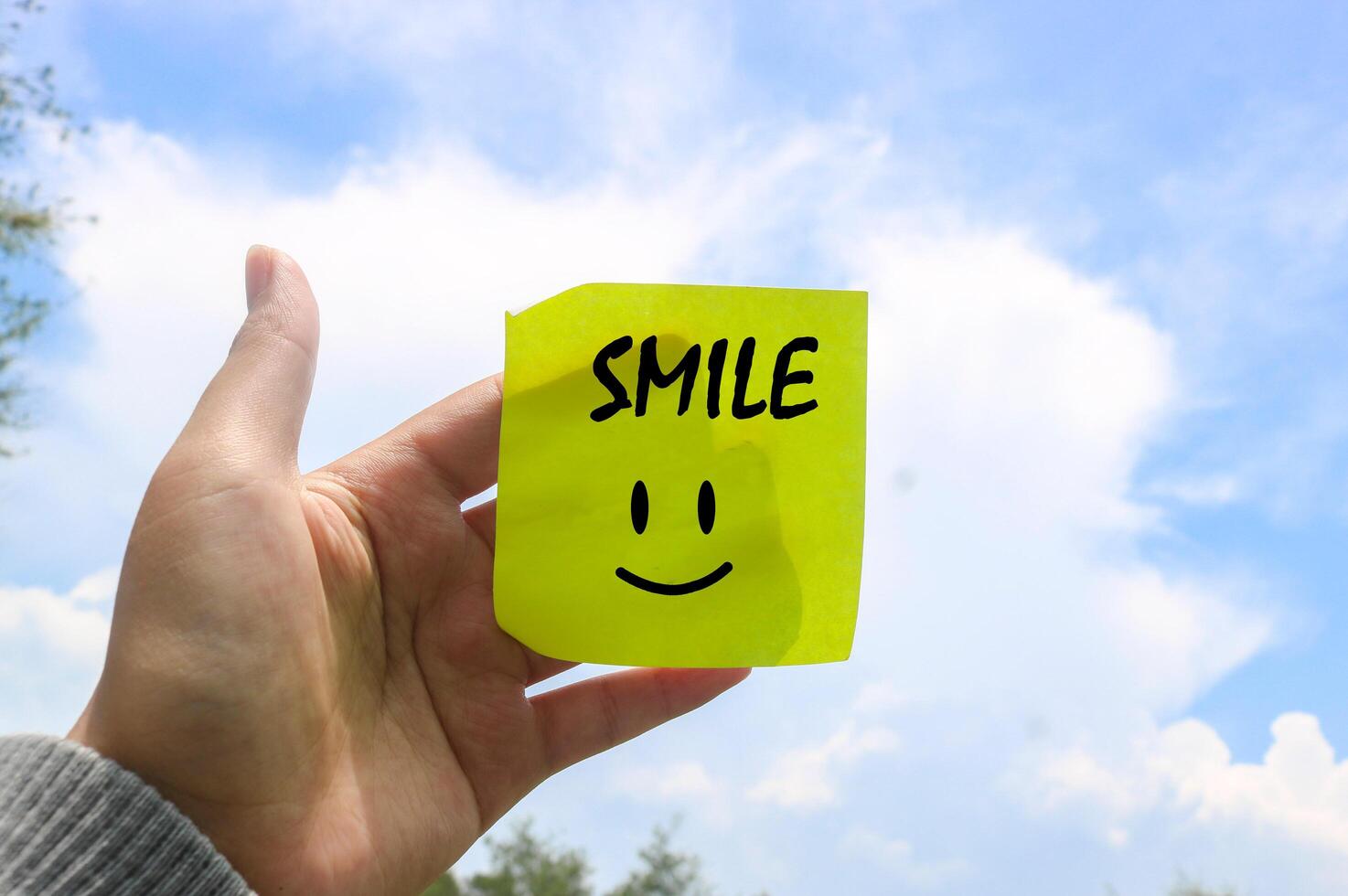 Hand holding note writing SMILE smile emoji with blue sky and clouds background. Motivational word wallpaper SMILE written on sticky note photo