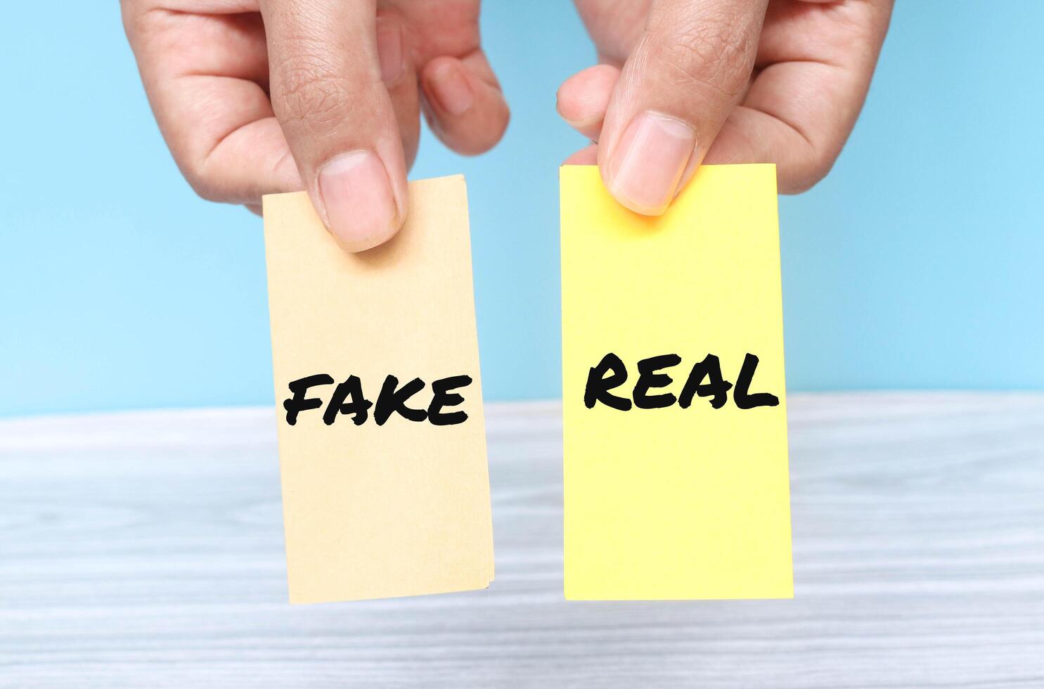 Hand holding the words Fake or Real on yellow shaped paper isolated on blue background. Real or fake business concept photo