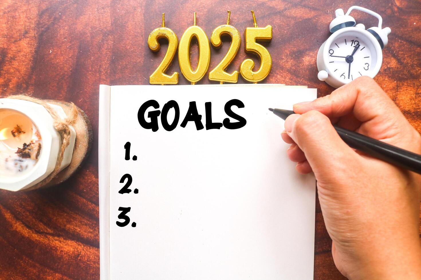 A person writes 2025 Goals in a notebook surrounded by aromatherapy candles and a clock. Planning a 2025 to do list photo