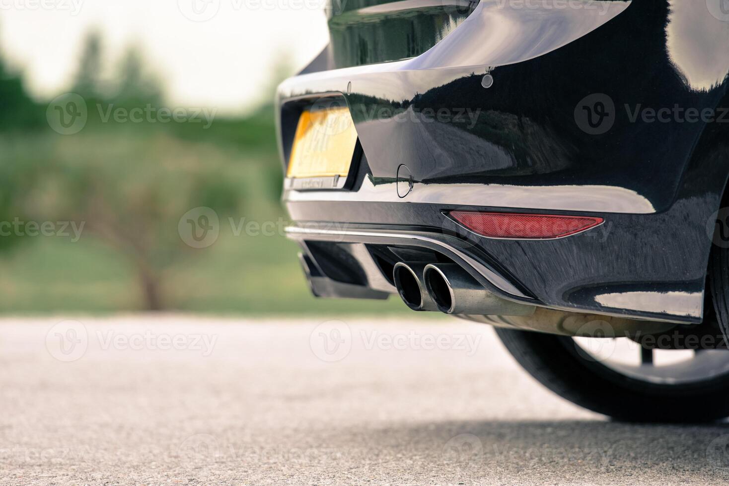 Close up of a black car's rear bumper and dual exhaust pipes The car is parked on a road with blurred greenery in the background photo