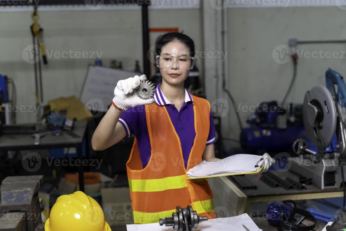Asian woman industrial engineer in vest hold a clipboard and looking check a example small gear at industrial workshop. photo