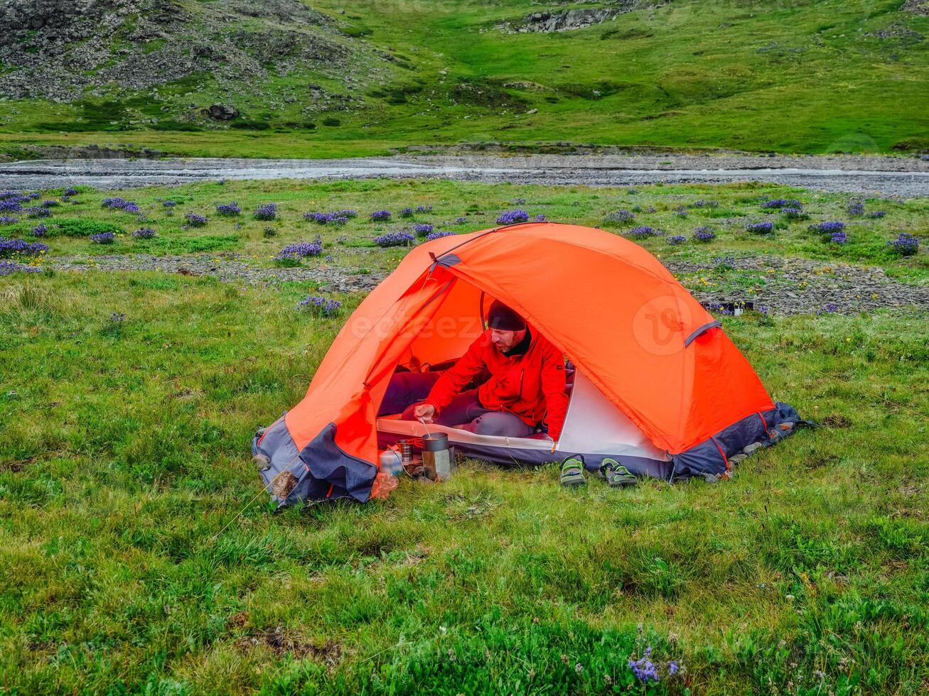 Camping on a summer green high-altitude plateau. Man in a orange tent is preparing food. Peace and relaxation in nature. photo