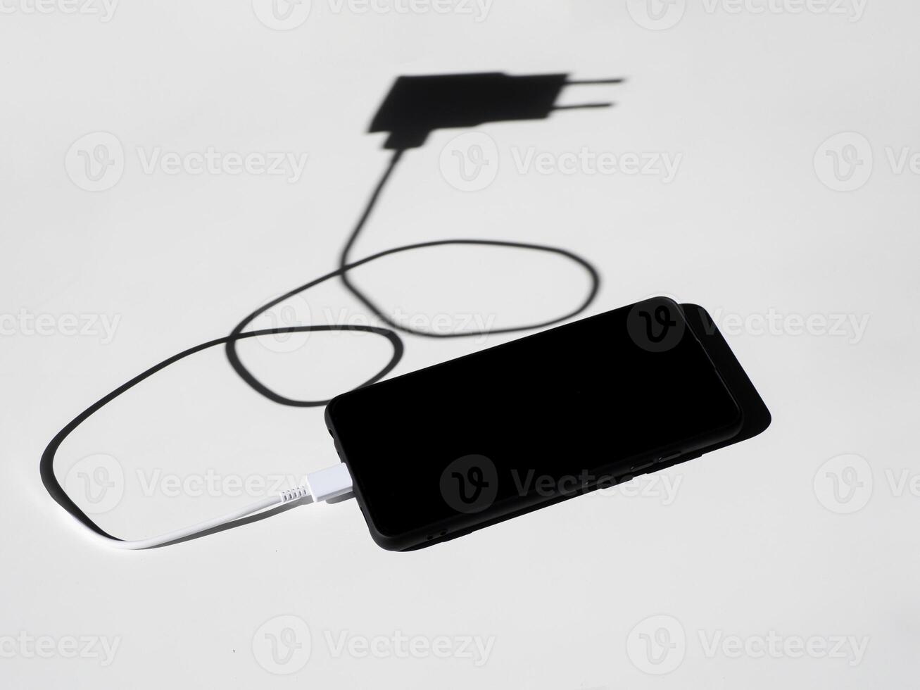 black smartphone with a charging wire on a white table with shadows. photo
