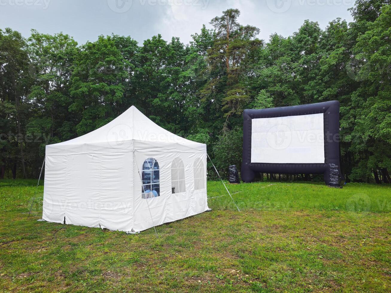 White small tent and large movie screen in the meadow at a park. photo