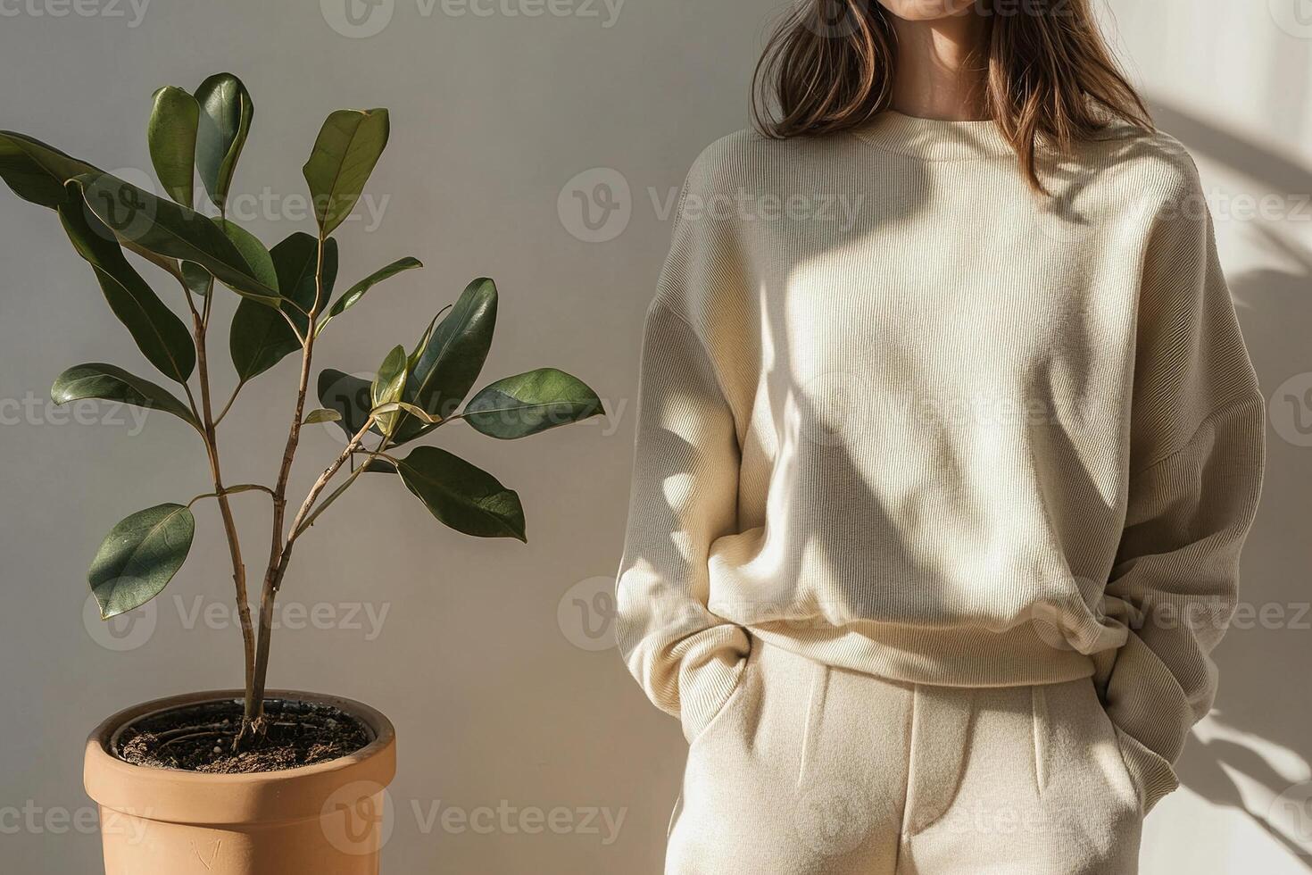A person stands in a sunlit indoor setting wearing a beige sweater and matching pants. A green houseplant in a terracotta pot adds a natural touch to the decor photo
