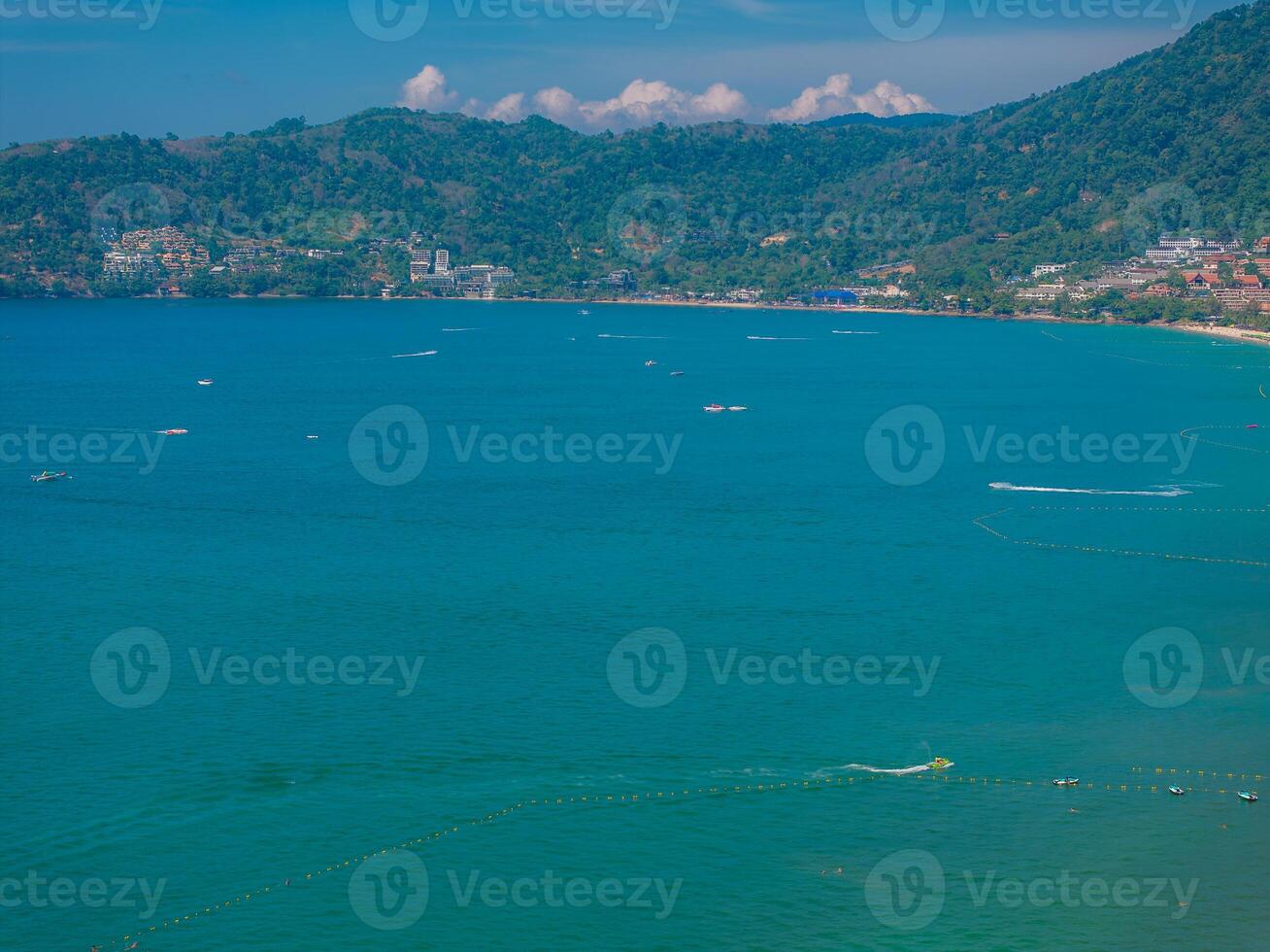 Aerial view of a turquoise bay in Phuket, Thailand, with small boats, lush green hills, scattered buildings, and a mountainous backdrop under clear skies. photo