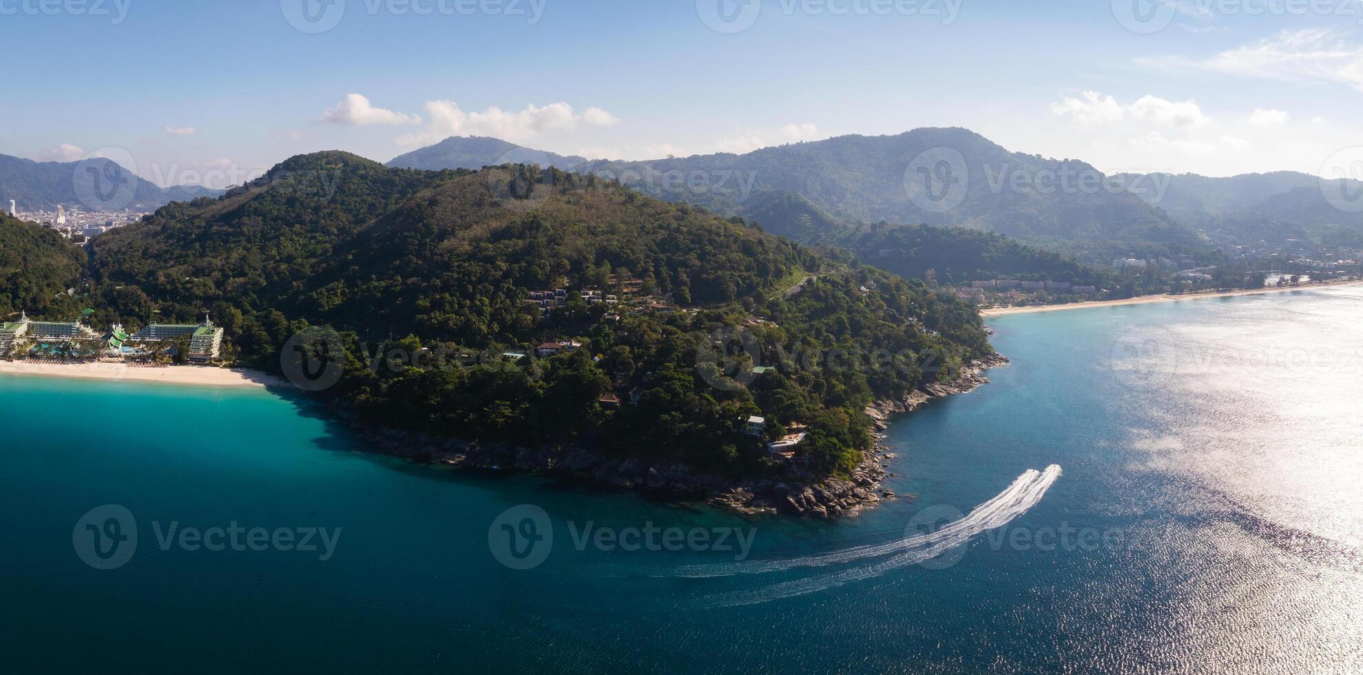 Aerial view of a green peninsula in Phuket, Thailand, surrounded by turquoise waters. A speedboat leaves a trail near a white sandy beach under a sunny sky. photo
