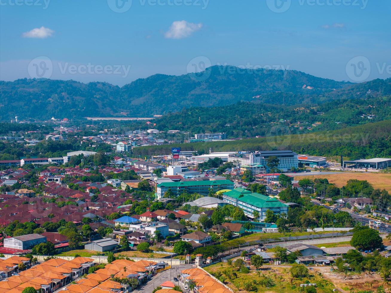 Aerial view of a town in Phuket, Thailand, featuring red tiled roofs, green hills, modern buildings, and a clear blue sky in the background. photo
