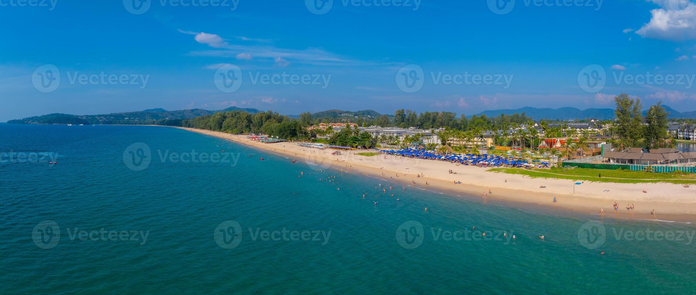 Aerial view of a long sandy beach in Phuket, Thailand, with turquoise waters, lush greenery, resort buildings, and rows of blue umbrellas along the shore. photo