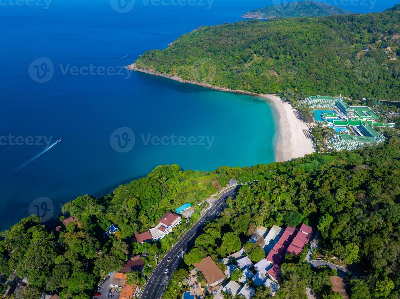 Aerial view of a crescent shaped beach in Phuket, Thailand, with turquoise waters, lush forest, colorful buildings, and a resort near the shoreline. photo