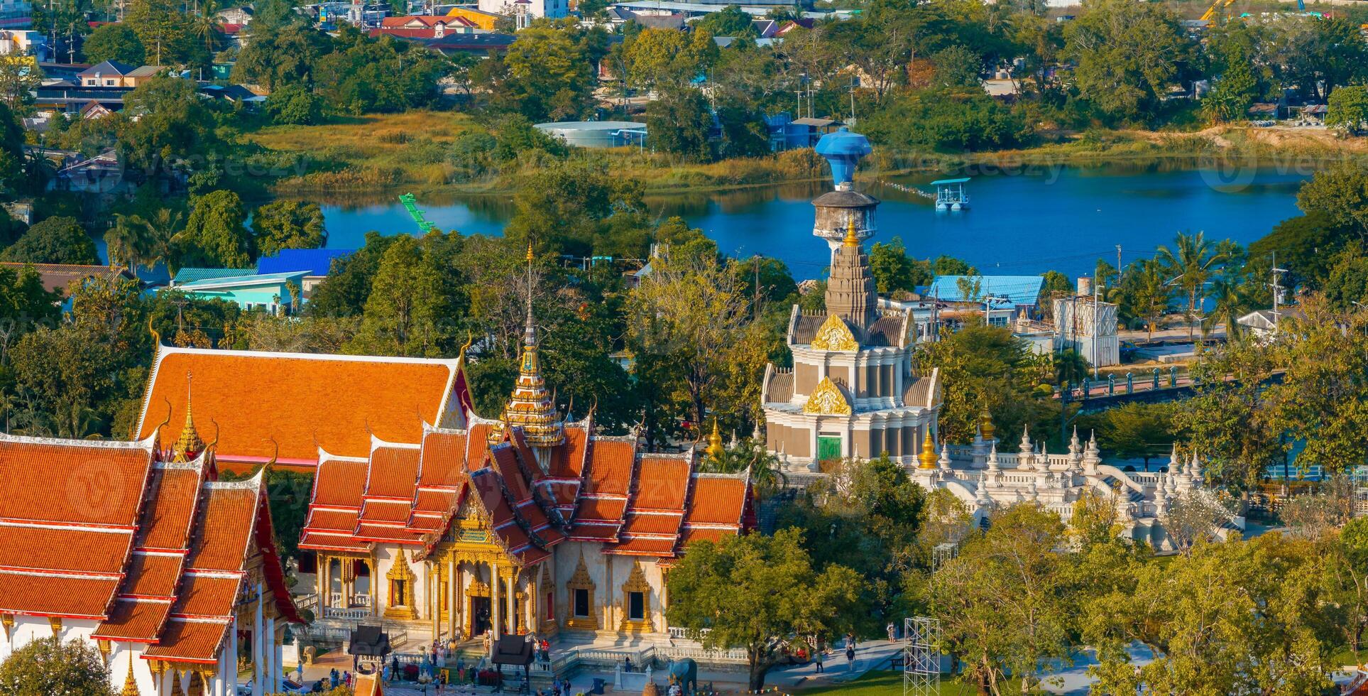 Aerial view of a temple complex in Phuket, Thailand, with ornate red and gold roofs, a blue domed structure, and a lotus pond surrounded by greenery. photo