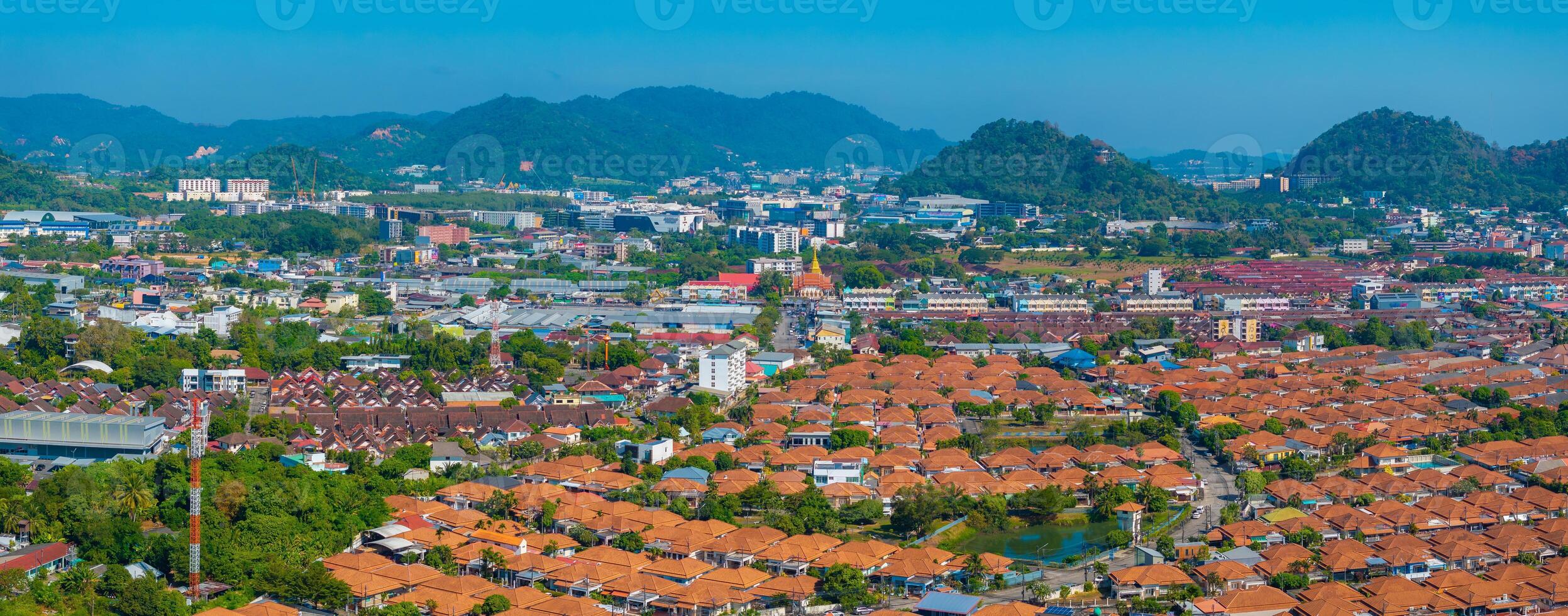 Aerial view of a vibrant urban area in Phuket, Thailand, featuring orange tiled roofs, a golden temple spire, and forested hills under a clear blue sky. photo