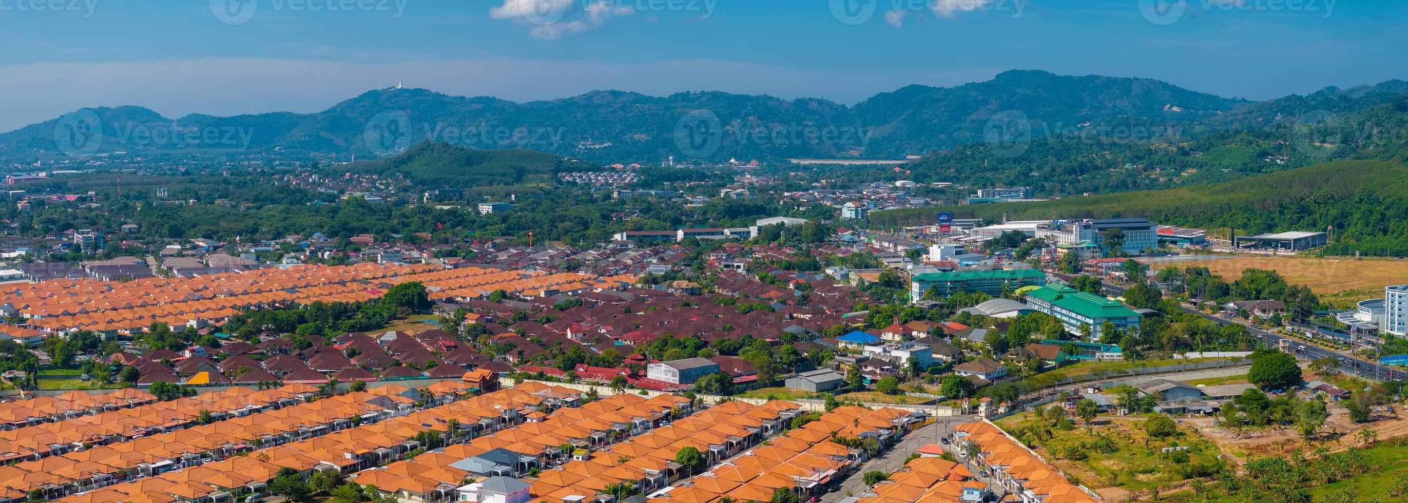 Aerial view of orange roofed houses in Phuket, Thailand, with lush green hills, the Big Buddha statue, scattered buildings, and a clear blue sky. photo