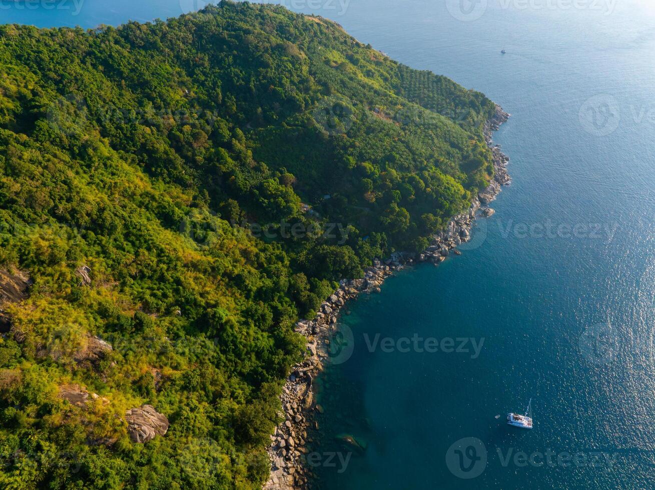 Aerial view of a lush, green coastline with rocky shores, clear blue waters, and a single white yacht near Phuket, Thailand's hilly terrain. photo