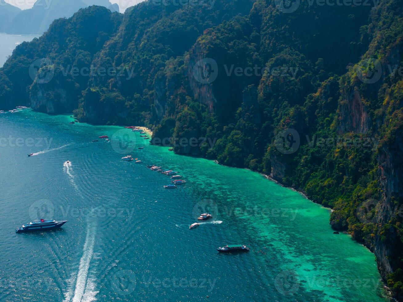 Aerial view of Phi Phi Islands in Thailand, showing turquoise waters, forested limestone cliffs, and boats leaving trails across the sea. photo