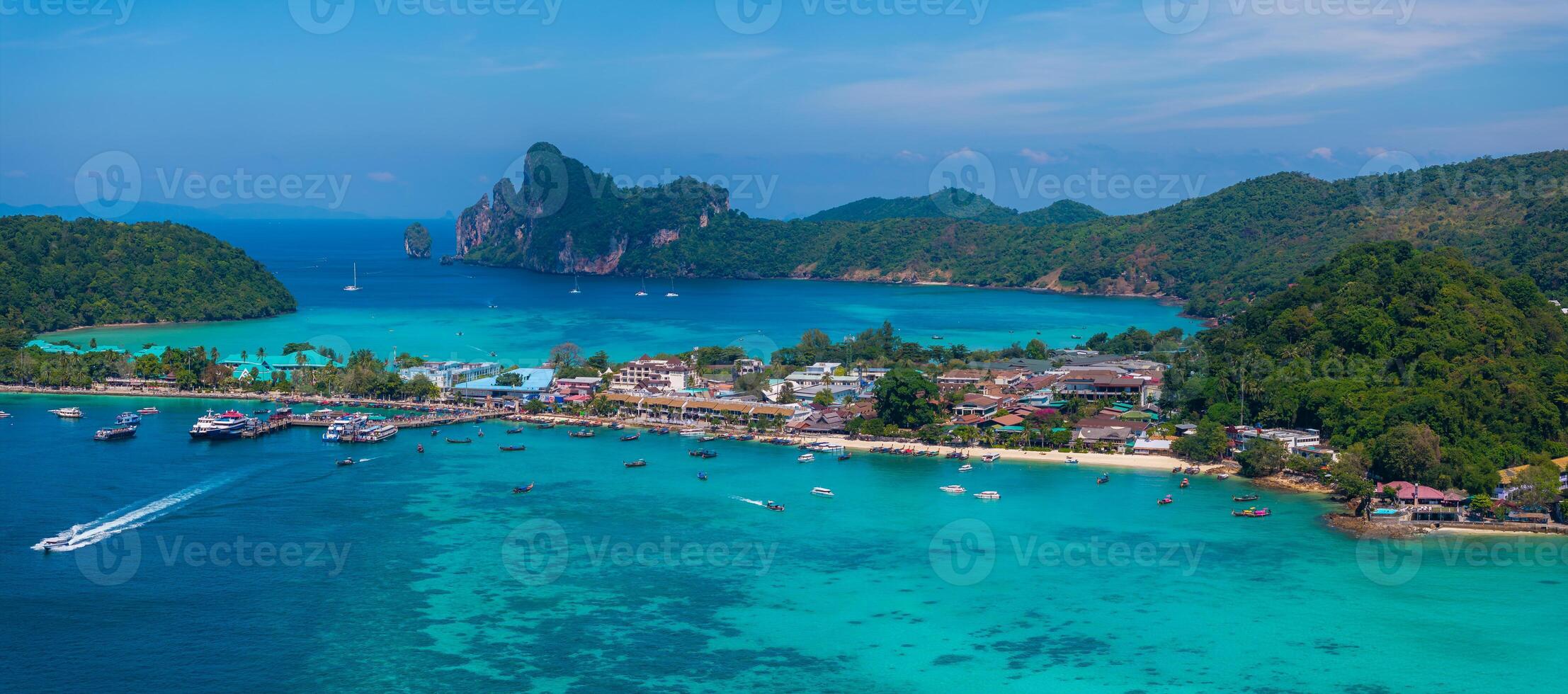 Aerial view of Phi Phi Islands in Thailand, showcasing turquoise waters, lush green hills, limestone cliffs, and a bustling coastal area with boats. photo