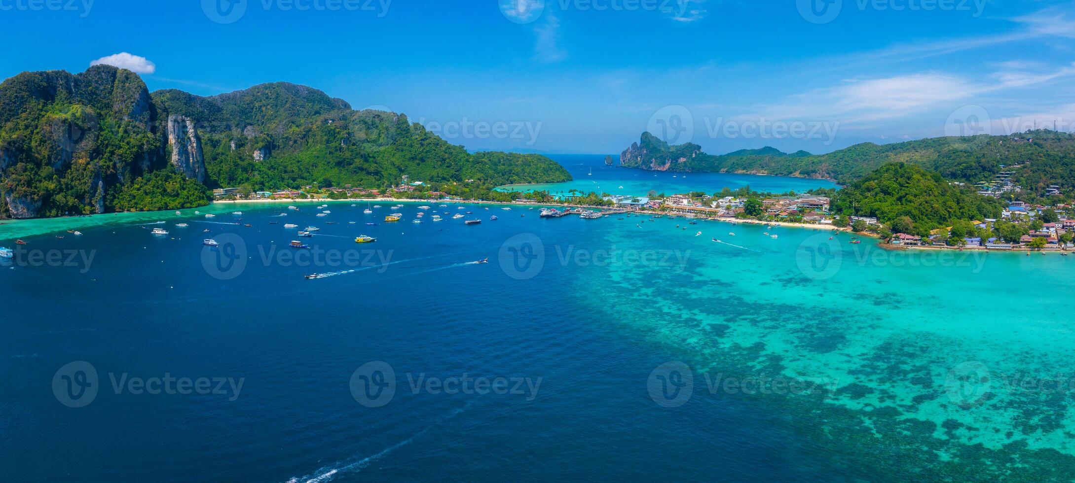 Aerial view of Phi Phi Islands in Thailand, showing turquoise waters, lush green hills, limestone cliffs, scattered boats, and coastal structures. photo