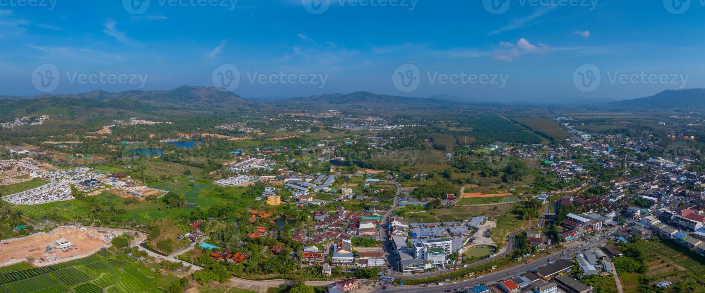 Aerial view of Phuket, Thailand, showing buildings, green fields, a lotus pond, rolling hills, and mountains under a clear blue sky. photo