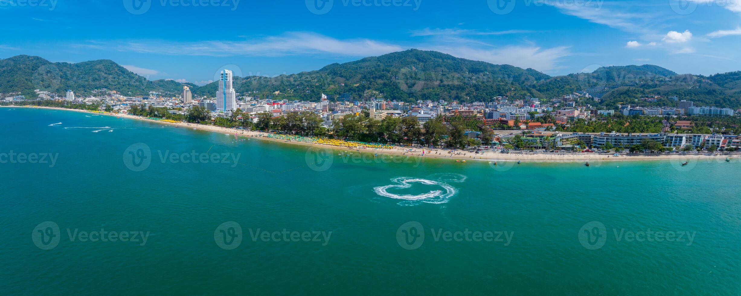 Aerial view of Phuket, Thailand, featuring a sandy beach, turquoise waters with circular patterns, lush green hills, and urban structures along the coast. photo