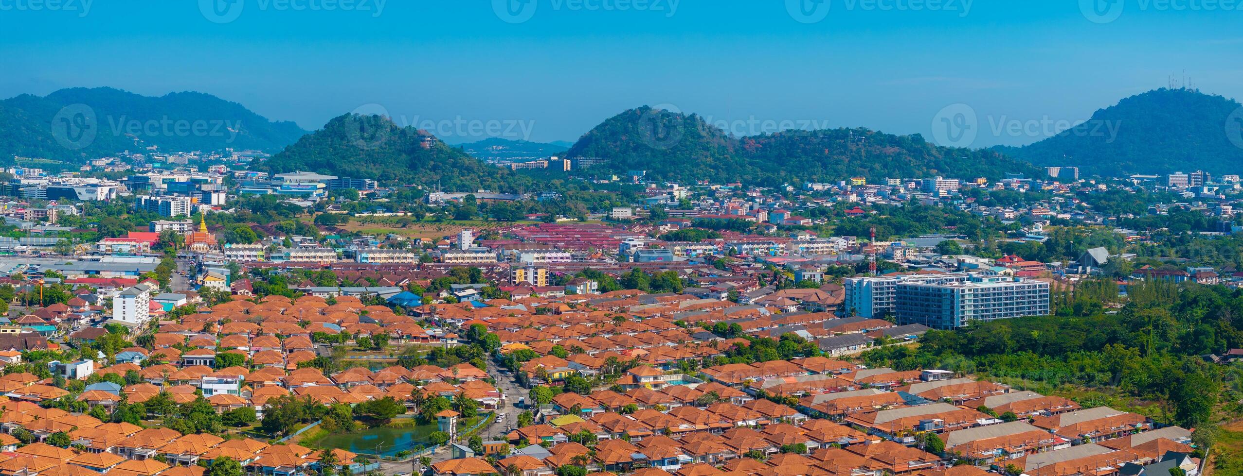 Aerial view of Phuket, Thailand, featuring orange roofed buildings, Wat Chalong's golden spire, and lush green hills under a clear blue sky. photo