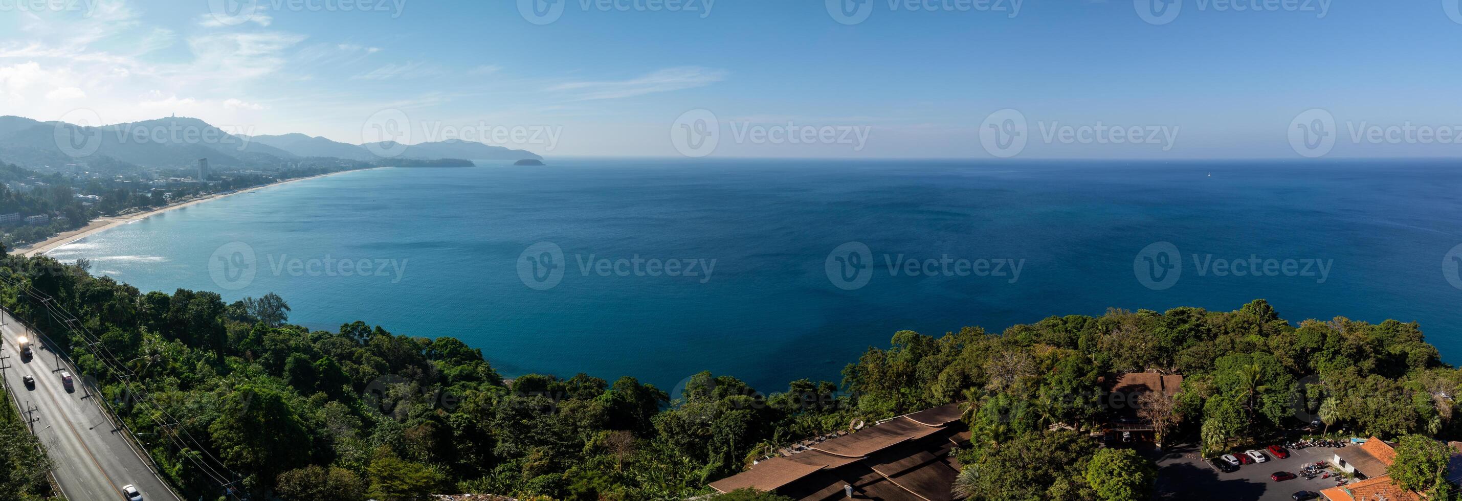 Aerial view of Phuket, Thailand, showing a calm blue ocean, green coastline, winding road, scattered buildings, and distant hills under a sunny sky. photo