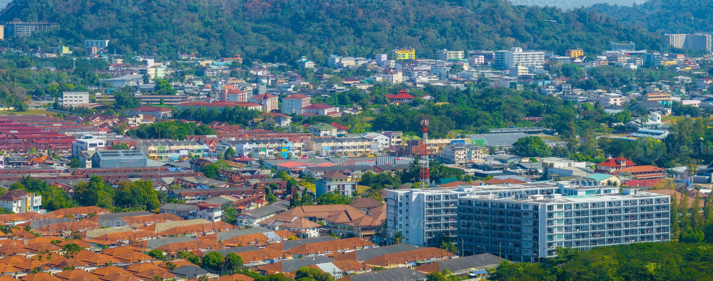 Aerial view of a bustling urban area in Phuket, Thailand, featuring modern buildings, red roofed houses, and lush green hills in the background. photo