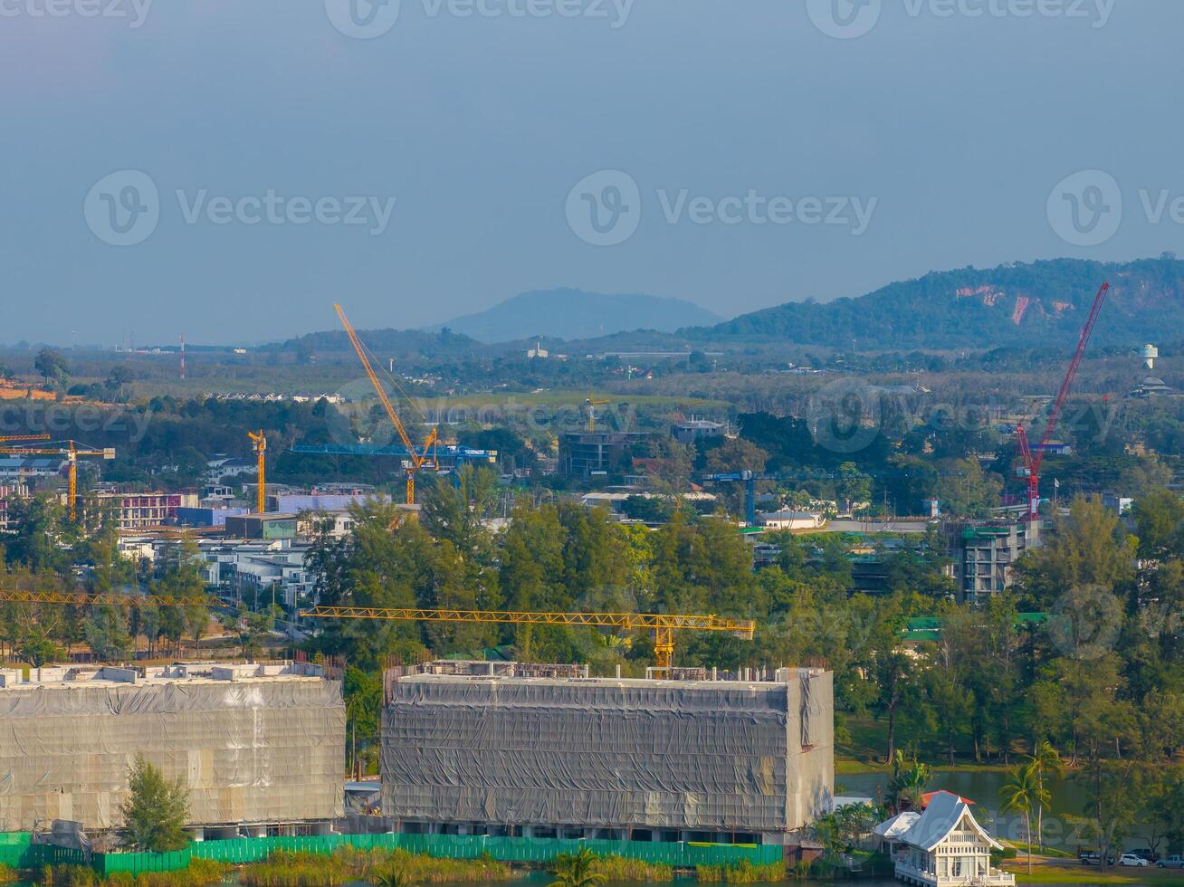 A construction site with cranes and scaffolding surrounded by dense vegetation and rolling hills in Phuket, Thailand. A small white building is in the foreground. photo