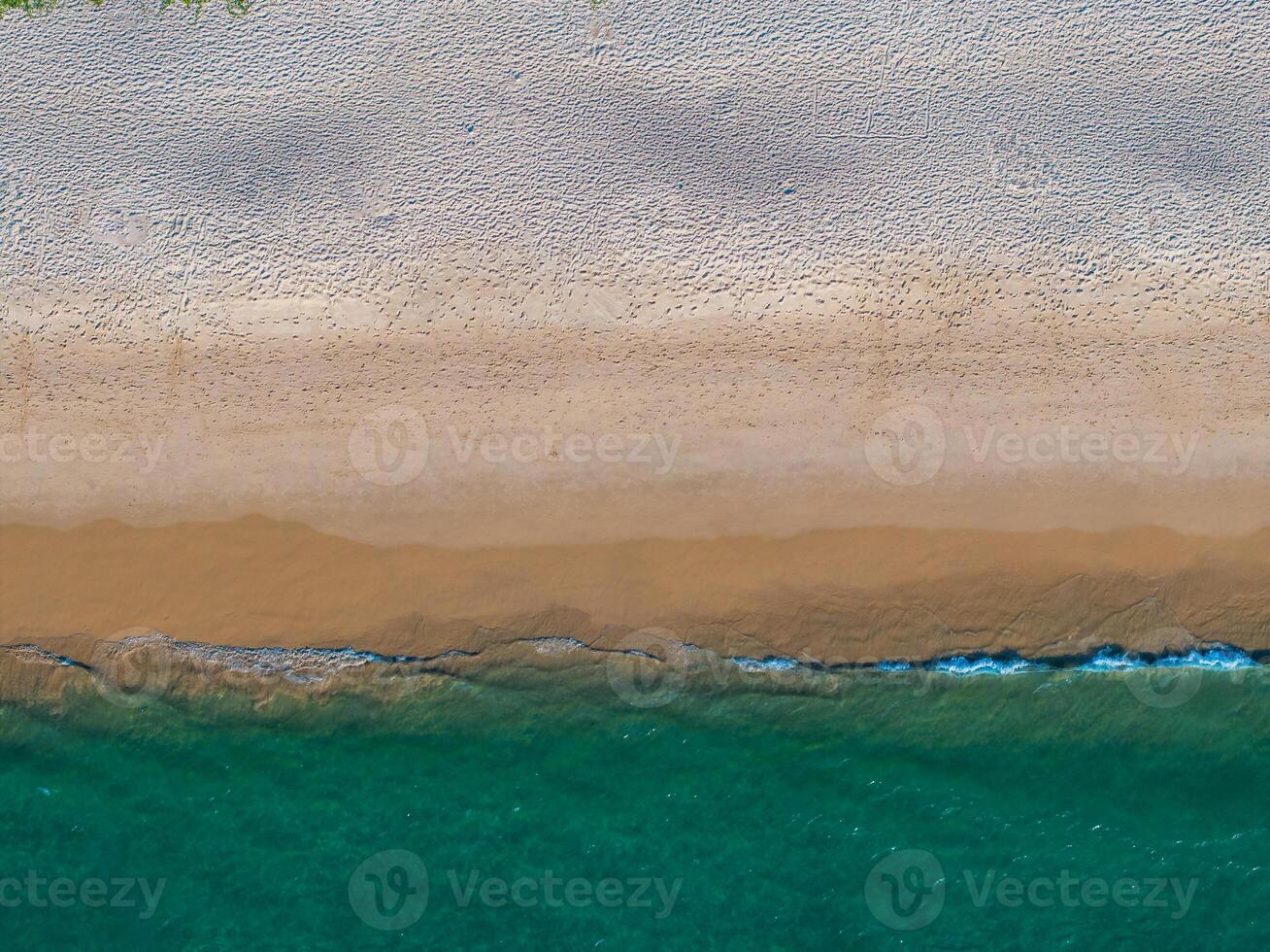 Aerial image of a beach with turquoise ocean waters, wet sandy shoreline, and dry textured sand. Sparse greenery is visible at the edge of the frame. photo