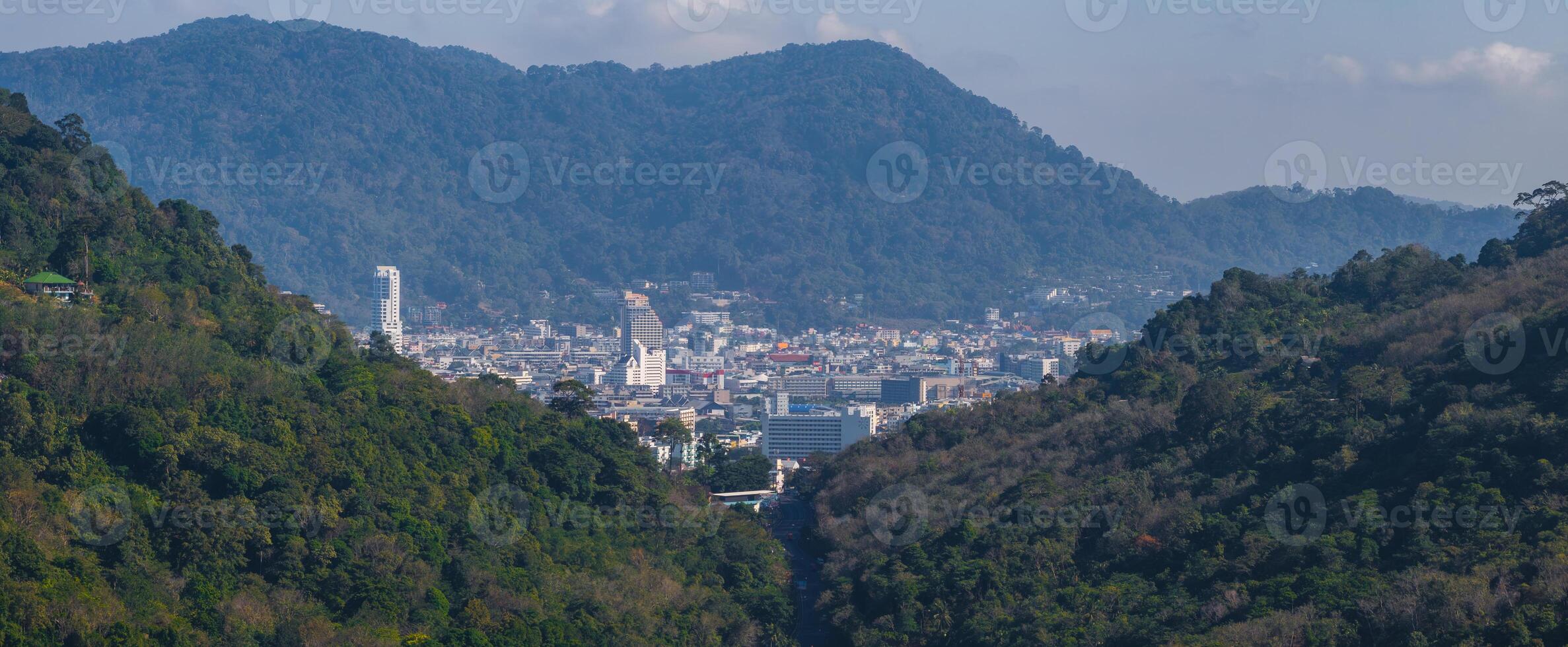 A city in Phuket, Thailand, surrounded by lush green hills and forested mountains. Prominent buildings include a tall white structure and a triangular one. photo