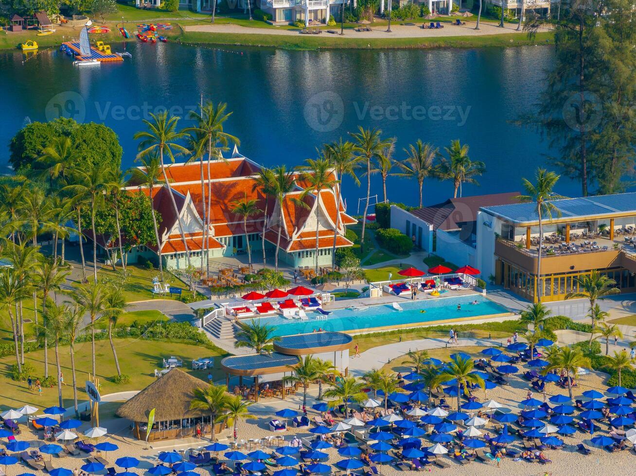 A vibrant beachfront resort in Phuket, Thailand, featuring a red roofed Thai style building, palm trees, a pool, beach umbrellas, and a serene lagoon. photo