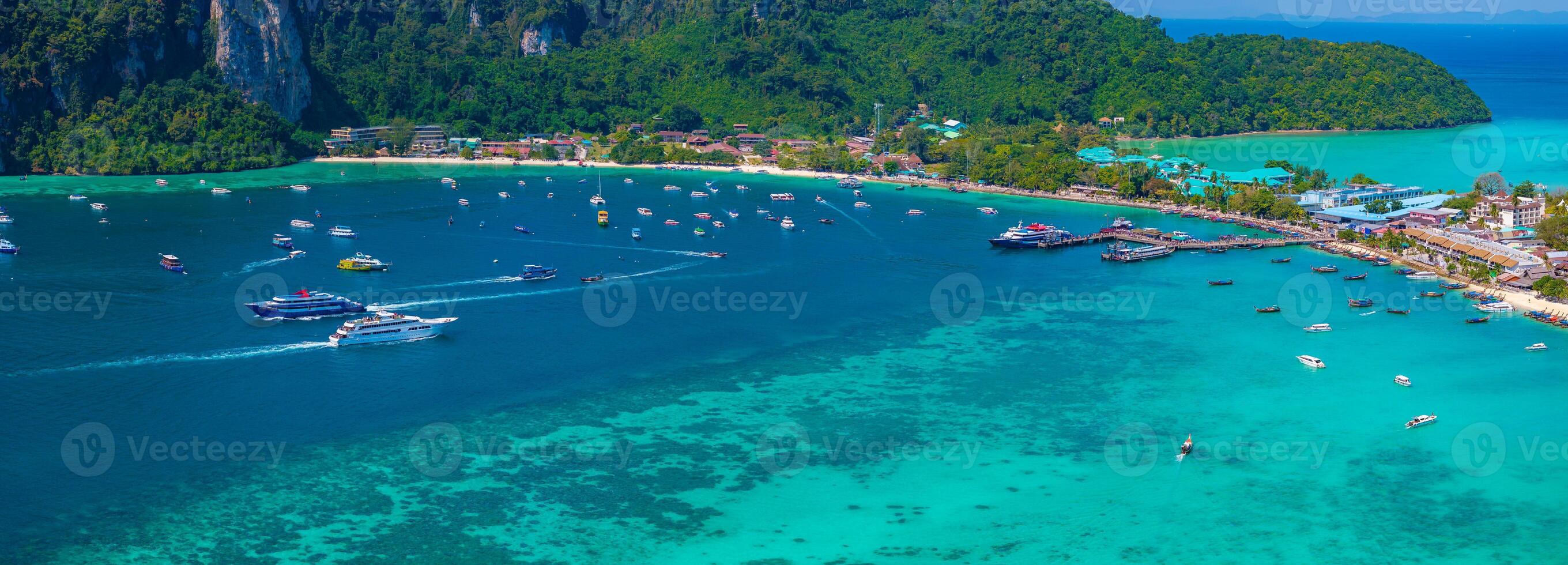 A vibrant aerial view of a bay in Phi Phi Islands, Thailand, featuring turquoise waters, boats, limestone cliffs, green hills, and developed shoreline areas. photo