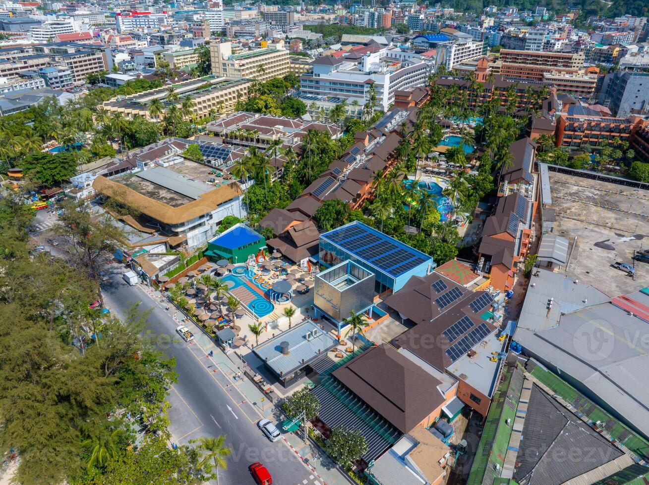 Aerial image of a bustling urban area in Phuket, Thailand, featuring a blue roofed building with a pool, surrounded by greenery, roads, and mixed architecture. photo