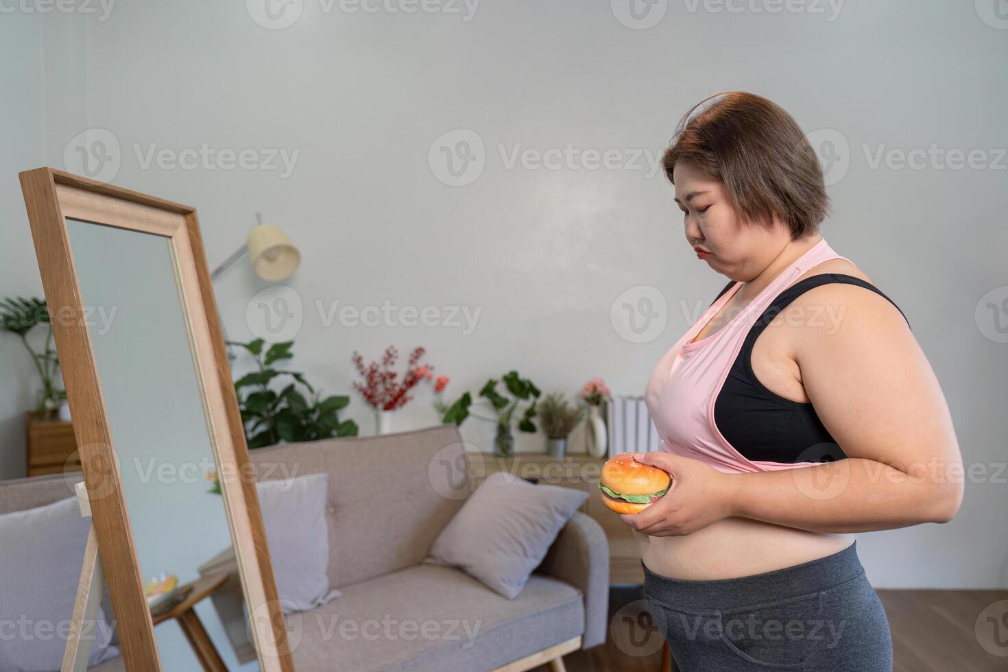Pensive woman holding a burger, gazing at her reflection in a mirror, contemplating her body image and choices photo