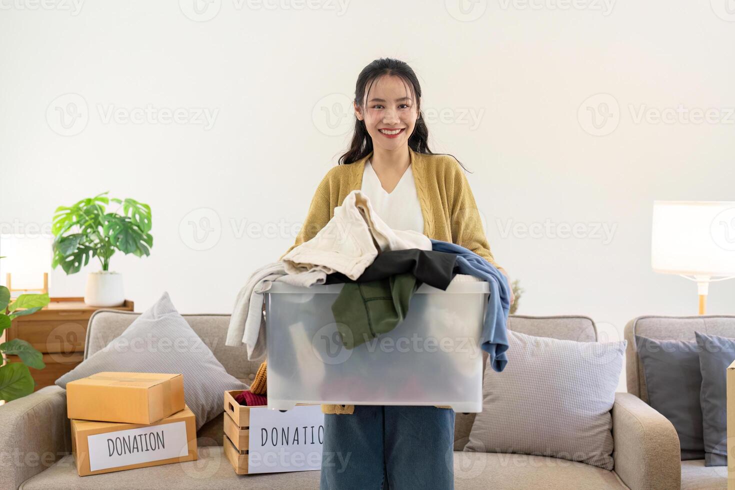 Smiling woman holding a transparent box full of clothes for donation, standing in a cozy living room photo