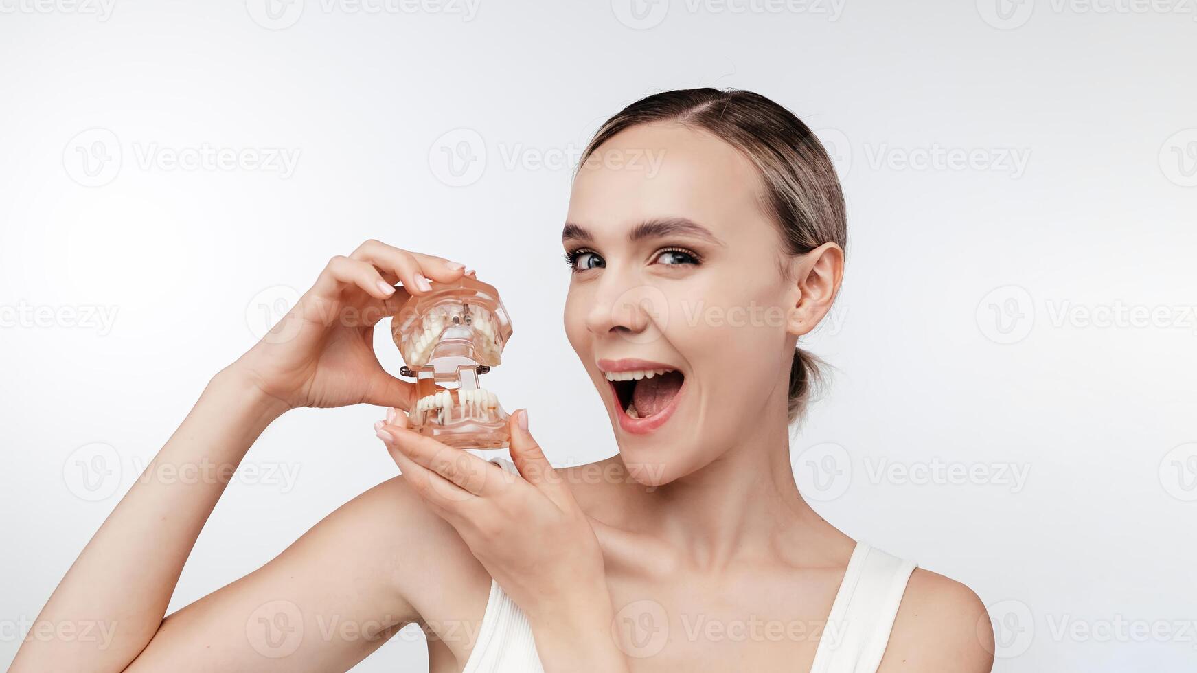 Happy young woman showcases a jaw model with dental implants. The concept of dentistry, dental health and veneers. Perfect for promoting dental services. Isolated on white studio background, free copy photo